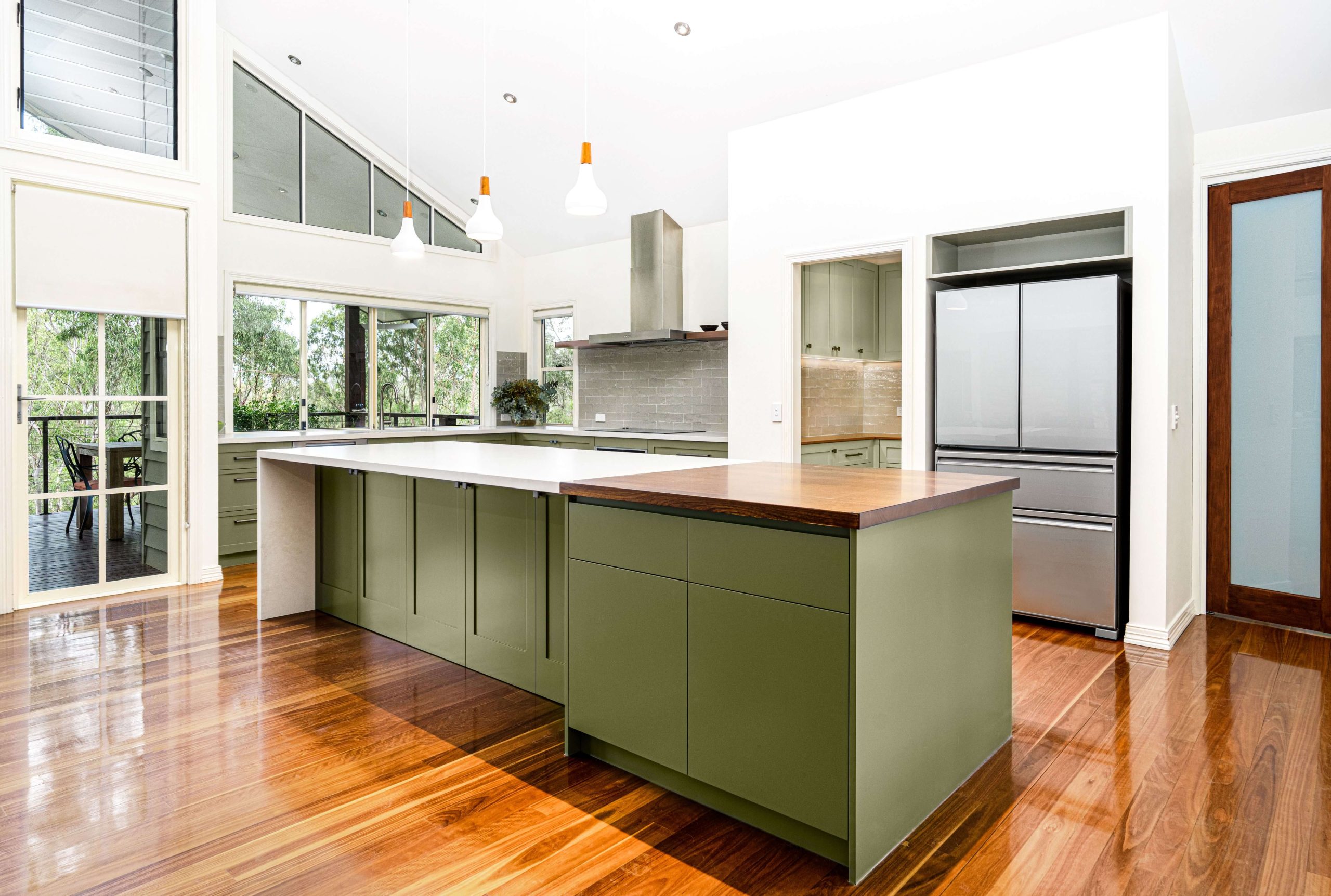 Contemporary kitchen remodel in Brisbane showcasing a spacious layout with olive green cabinets and a large central island. The island features a white countertop with a wooden extension, complementing the polished hardwood floors. Ample natural light streams through large windows and glass doors, enhancing the modern design.