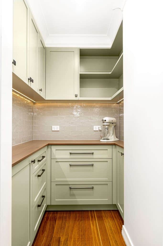 Compact pantry area in a modern Brisbane kitchen remodel featuring olive green cabinets and drawers with black handles. The space includes white countertops, a sleek subway tile backsplash, and polished hardwood floors. A built-in shelf offers additional storage, and under-cabinet lighting enhances the clean, stylish design. An electric mixer is neatly placed on the counter.