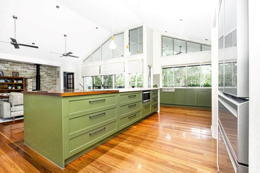 Spacious Brisbane kitchen remodel featuring olive green cabinetry, a large central island with wooden countertop, and polished hardwood floors. The design includes modern appliances, large windows providing natural light, and pendant lighting. The open-plan layout connects to a cozy living area with stone wall accents and ceiling fans, creating a seamless flow between spaces.