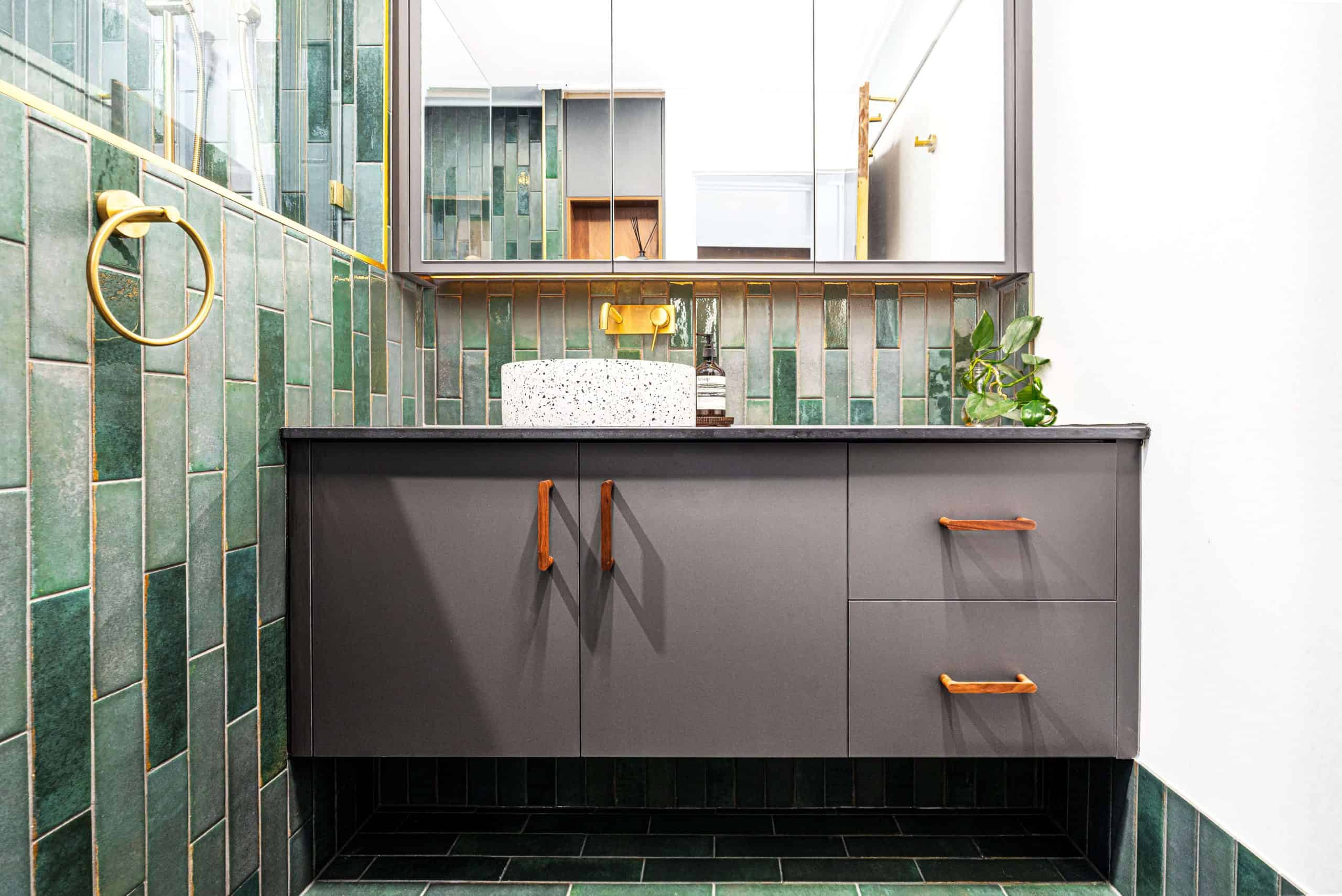 Modern Brisbane bathroom featuring green subway tiles, a floating vanity with dark grey cabinets and wooden handles, and a terrazzo vessel sink on a grey countertop. The design includes gold fixtures, a mirrored cabinet, and a touch of greenery, creating a sleek and contemporary aesthetic.