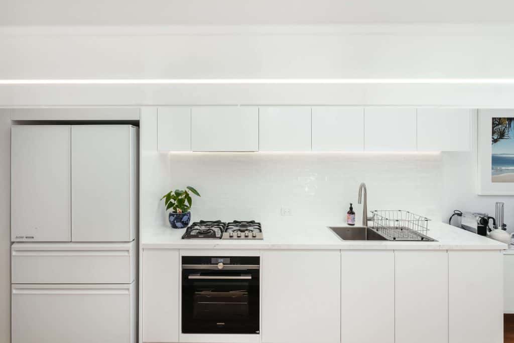 Modern and well-lit kitchen space in New Farm, Brisbane, featuring under-cabinet LED lighting, white subway tiles, and stainless steel appliances, with a framed coastal landscape picture adding a serene touch.