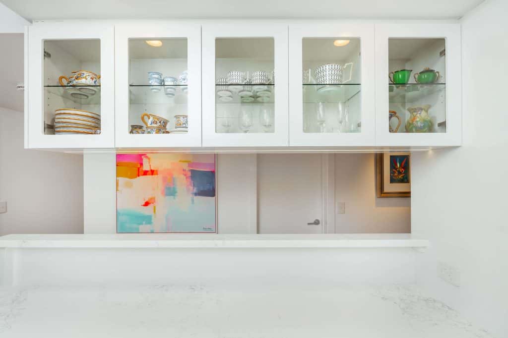 Brightly lit display cabinets in a New Farm, Brisbane kitchen remodel, showcasing a collection of colorful and patterned crockery against a backdrop of modern artwork and white countertops.