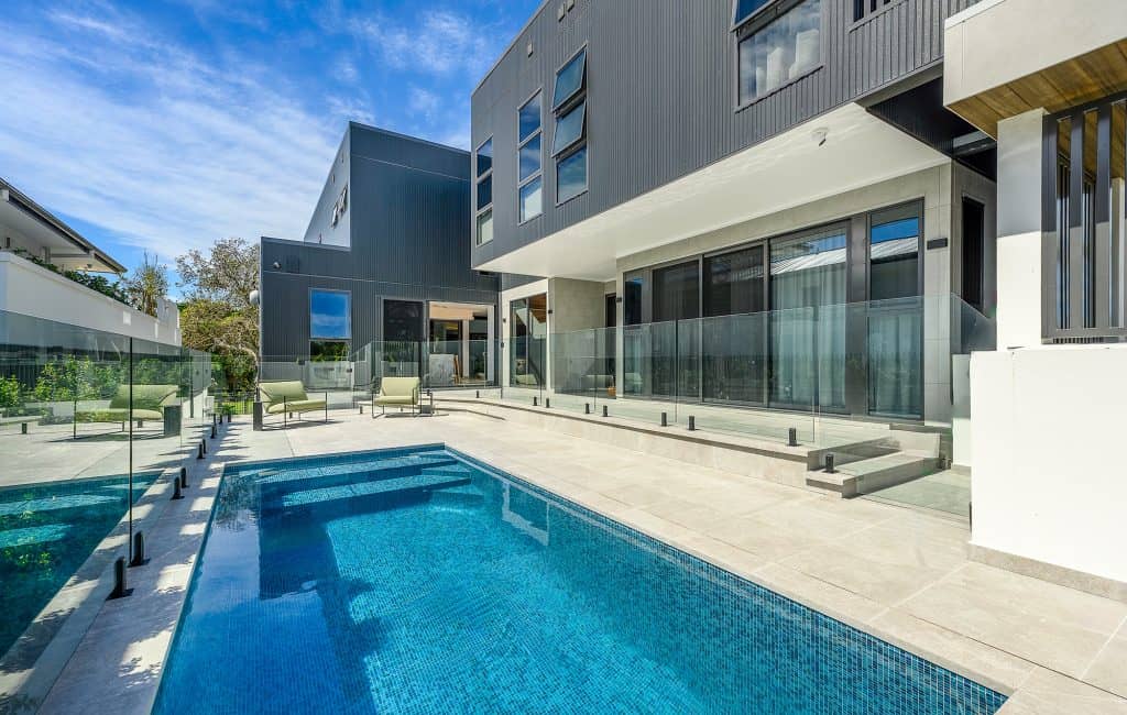 A modern backyard with a pristine swimming pool and surrounding glass fence, showcasing sleek architecture and contemporary design. The pool area is complemented by comfortable lounge chairs and a spacious patio, highlighting the luxurious outdoor living space of the Pottsville Project.