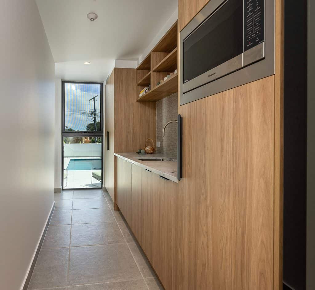 Modern butler's pantry featuring sleek wooden cabinetry, open shelving, and a built-in microwave. The space includes a small sink with a stylish faucet and offers ample storage for kitchen essentials. A large window at the end of the hallway provides natural light and a view of the outdoor pool area, enhancing the room's airy and functional design.