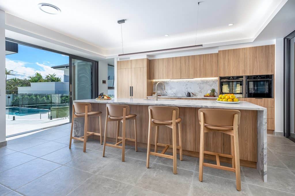 Modern kitchen interior featuring a large marble island with four wooden bar stools, sleek wood cabinetry, and integrated appliances. The open layout extends to a view of the outdoor pool area through large glass sliding doors, creating a seamless indoor-outdoor living space. Fresh fruit and decorative items add a touch of warmth and elegance to the sophisticated design.