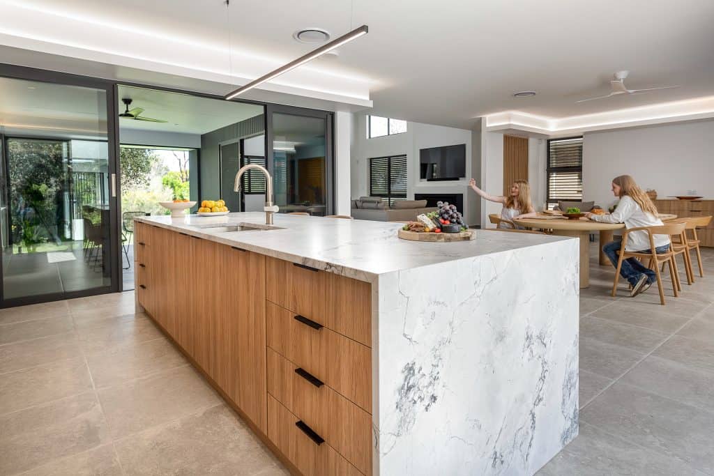 Spacious modern kitchen with a large marble island featuring wooden cabinetry and a sleek stainless steel faucet. The kitchen opens up to a bright dining area where two young girls are seated at a round wooden table. Large glass doors provide access to a lush outdoor space, creating a seamless flow between indoor and outdoor living. Decorative trays with fresh fruits and a light fixture add elegance to the contemporary design.