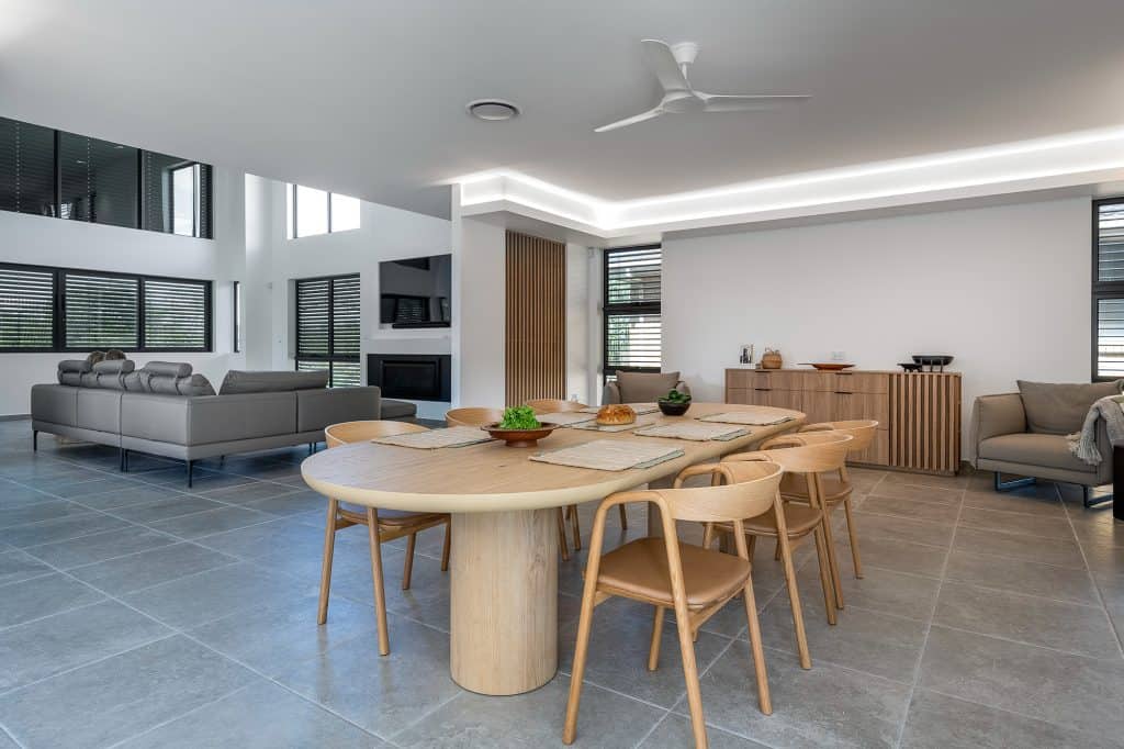 Spacious open-concept living and dining area featuring a large wooden dining table with matching chairs. The living room includes modern gray sofas and a wall-mounted television. The space is well-lit with natural light from large windows with black frames and enhanced by recessed ceiling lighting. A light wood sideboard and minimalist decor complete the contemporary design.