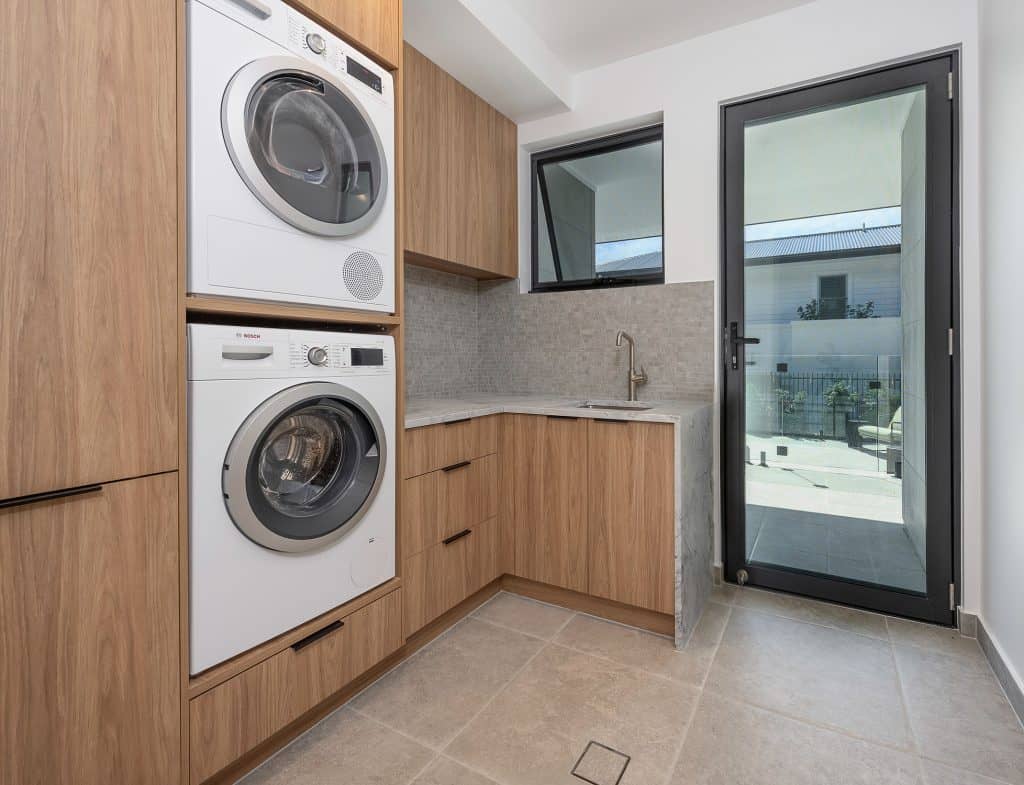 Modern laundry room with a stackable Bosch washer and dryer set integrated into custom light wood cabinetry. The room features a small sink with a marble countertop, a tiled backsplash, and a black-framed glass door leading to the outside. Natural light enters through a window above the sink, enhancing the clean and functional design.