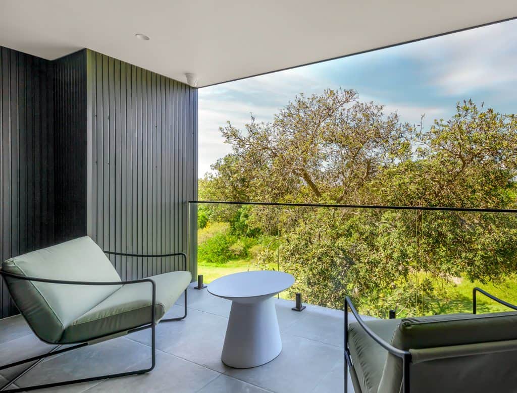 Modern outdoor balcony featuring sleek grey walls and flooring, with a minimalist design. The balcony is furnished with two comfortable chairs and a small white table, offering a serene spot to relax. The glass railing provides an unobstructed view of the lush green trees and surrounding nature, enhancing the tranquil ambiance of this outdoor space.