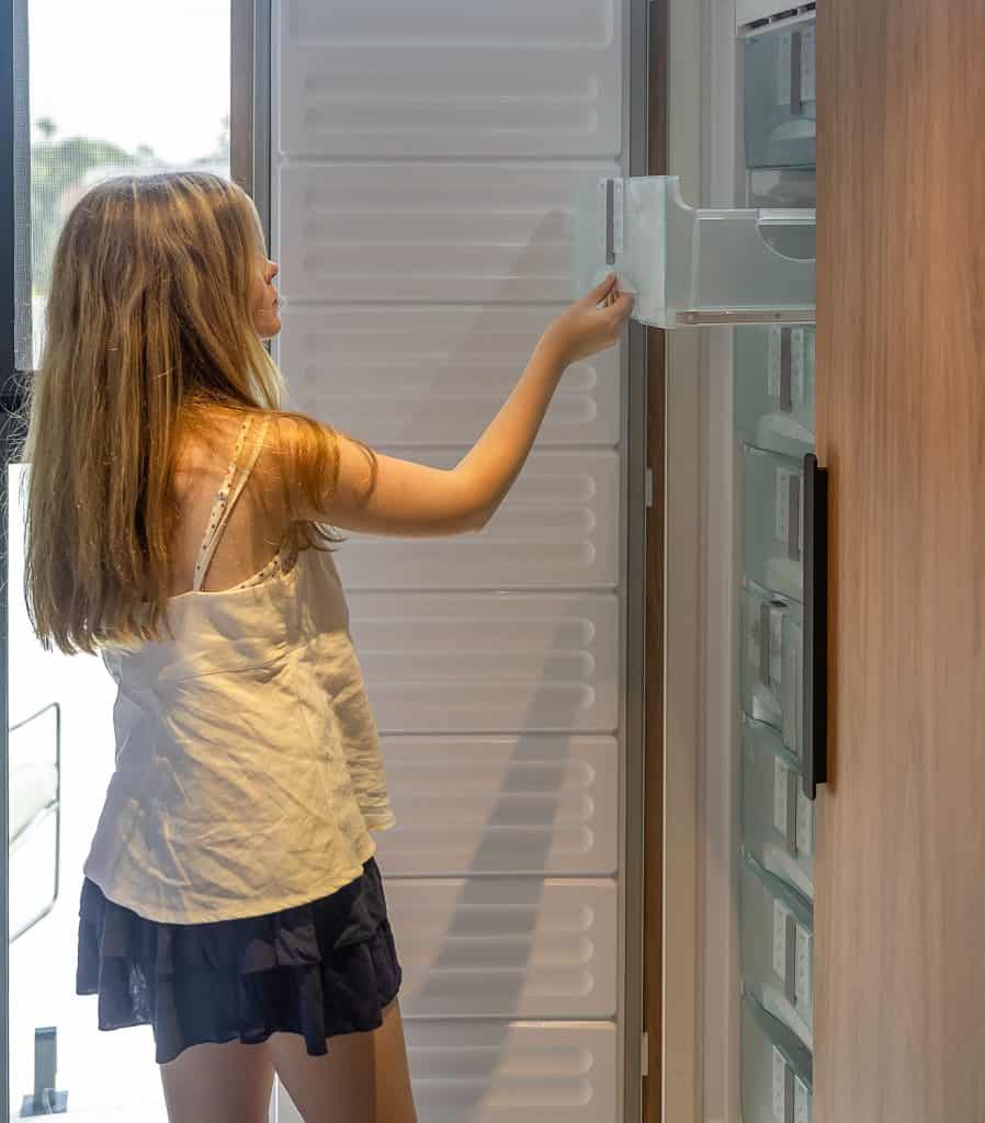 A young girl opening a drawer in a tall, integrated freezer unit. The appliance is built into custom light wood cabinetry, blending seamlessly with the kitchen design. Natural light from a nearby window illuminates the space, highlighting the modern and functional kitchen layout.