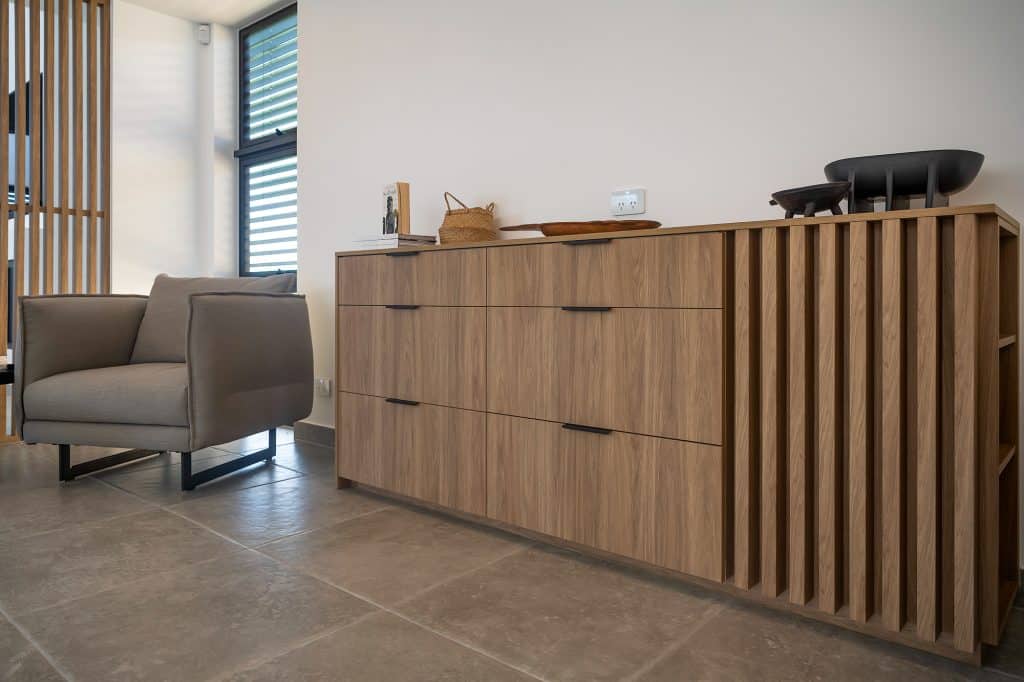 Modern living area featuring a sleek wooden buffet with black handles, showcasing a blend of drawers and slatted design elements. A comfortable grey armchair is positioned nearby, adding a touch of coziness to the space. Decorative items and books are arranged on the buffet, enhancing the room's contemporary and inviting atmosphere.