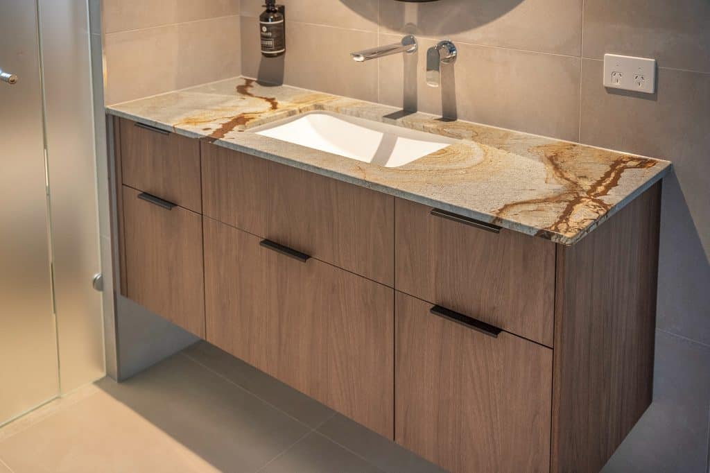 Modern bathroom vanity featuring a unique marble countertop with natural veining and integrated sink. The wood-toned cabinetry offers ample storage with sleek black handles, enhancing the contemporary design. A wall-mounted faucet and a soap dispenser complement the elegant and functional setup, reflecting the high-quality craftsmanship of the Pottsville Project.