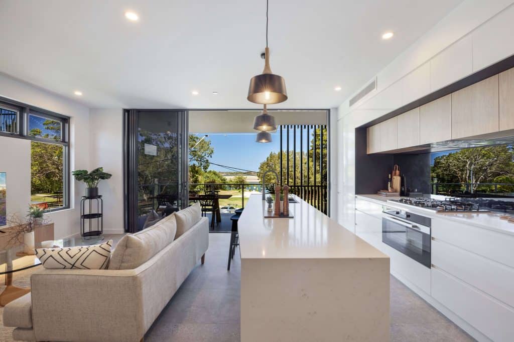 Modern kitchen with a sleek white waterfall island countertop, stainless steel appliances, and wood-finish cabinets opening up to a cozy living area with a view of the outdoor patio.