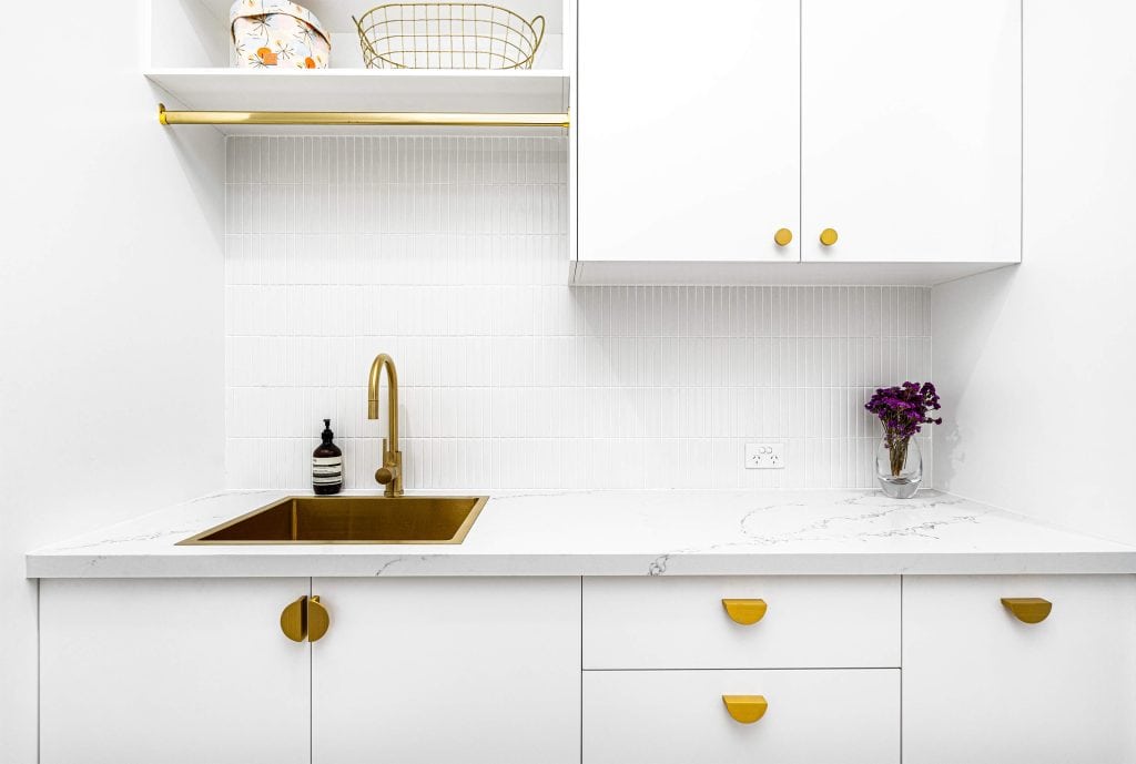 Close-up of a laundry room sink area with a sleek gold faucet, white subway tile wall, and white cabinets featuring distinctive gold pulls, set against a marble-like countertop.