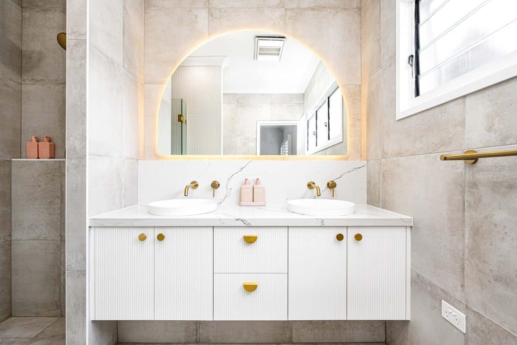 Spacious double vanity with two round basins, gold faucets, under-cabinet lighting, and a large arched mirror with a golden glow