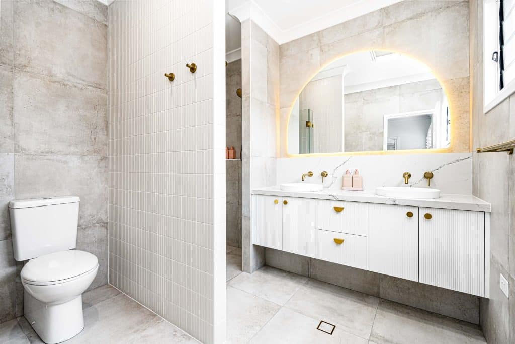 Bright bathroom with white tiled walls, a toilet, and a vanity with dual basins, gold fixtures, and a softly lit large arch mirror.